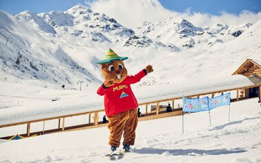 Maskottchen Murmli auf Skiern in der Kinderschneealm im Hintergrund der überdachte Förderbandtunnel Blog Serfaus-Fiss-Ladis in Tirol | © Skischule Serfaus